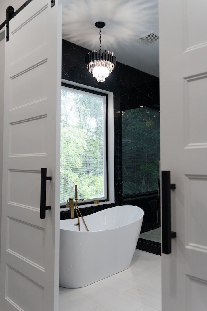 Open doors reveal a sleek bathroom with a white freestanding tub, black marble walls, chandelier, and a large window to green trees. Architecture by James Knight. Photography by Kalyn Kight