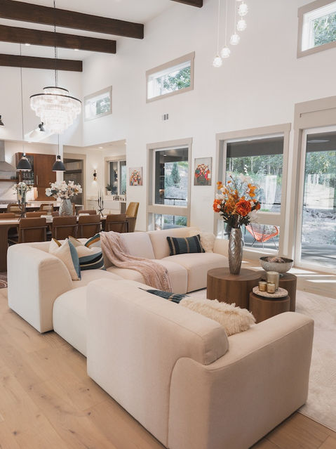 Bright living room area featuring a large cream modular sectional sofa, nested round wooden coffee tables, light hardwood floors, and large windows looking out to a wooded landscape. Architecture by James Knight Photo by Kalyn Kight Reynard Architectural Designs