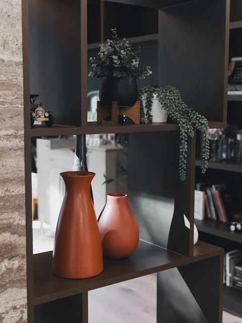 Close-up view of an asymmetrical dark wood shelving unit decorated with two smooth terracotta vases, a small vintage toy car, and potted green plants. Architecture by James Knight Photo by Kalyn Kight Reynard Architectural Designs