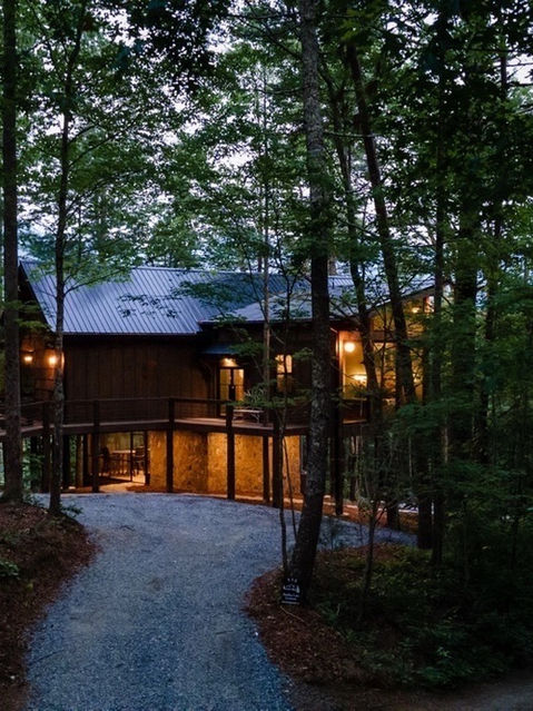 A wide exterior view of a modern, multi-level wooden cabin surrounded by dense, tall trees at dusk. Warm light glows from the interior windows and under-deck area, with a curving gravel driveway leading up to the property.
Architecture by James Knight Photography by Kalyn Kight. Reynard Architectural Designs