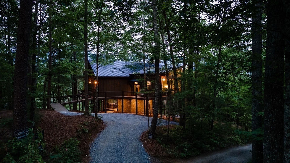 Exterior view at twilight of a modern rustic treehouse cabin nestled among tall trees, featuring dark wood siding, a metal roof, warm glowing interior lights, and an elevated wooden walkway accessed by a gravel driveway. A gold badge in the bottom right corner reads "Atlanta Magazine's HOME Georgia Design Awards GOLD WINNER 2023.