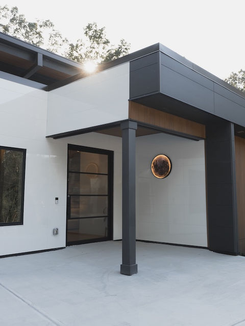 Close up of a modern home exterior featuring white paneling, dark gray trim, warm wood siding, a glass-paneled black front door, and a concrete patio. Architecture by James Knight Photo by Kalyn Kight Reynard Architectural Designs