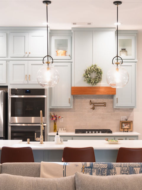 Kitchen interior with light blue cabinets, a white island with brown leather barstools, three large glass globe pendant lights, and stainless steel appliances. Custom home designed by Reynard Architectural Designs