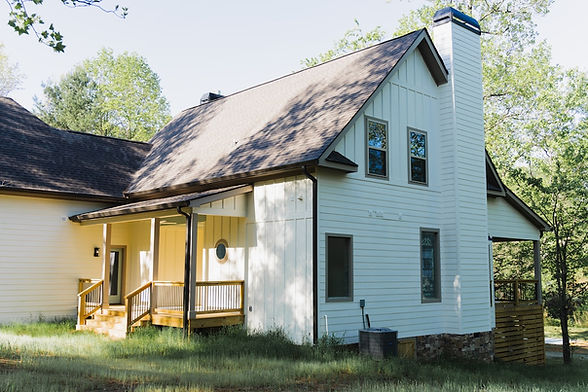 Side elevation of a custom modern mountain home in Hiawassee, GA, showcasing white board-and-batten siding, a large dark shingled roof, a covered wooden porch, and stone foundation accents. Reynard Architectural Designs