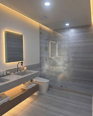 Thompson Creek- Bathroom. A modern bathroom featuring gray linear stone tiles on the walls and floor, a walk-in glass shower with steam rising, a wall-hung toilet, a long wood floating vanity with a concrete sink, a backlit rectangular mirror, and warm accent lighting throughout. Reynard Custom Homes architectural designs.