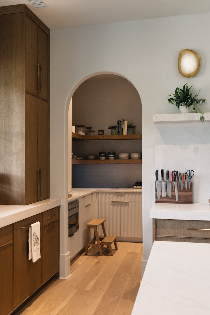 Modern kitchen with wood cabinets, marble countertops, knives on a stand, and a stool. Shelves with dishes and books in the background. Reynard Architectural Designs.