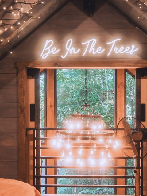A view from a lofted bedroom looking through a doorway with a neon sign that says 'Be In The Trees', showing a chandelier hanging over the lower living space, framed by branch lights. Architecture by James Knight Photography by Kalyn Kight. Reynard Architectural Designs