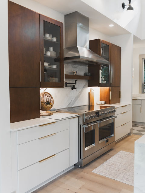 Detail shot of a modern kitchen showing a large stainless steel double oven range, matching vent hood, white lower cabinets with brass pulls, dark wood upper cabinets, and a marble backsplash. Architecture by James Knight Photo by Kalyn Kight Reynard Architectural Designs