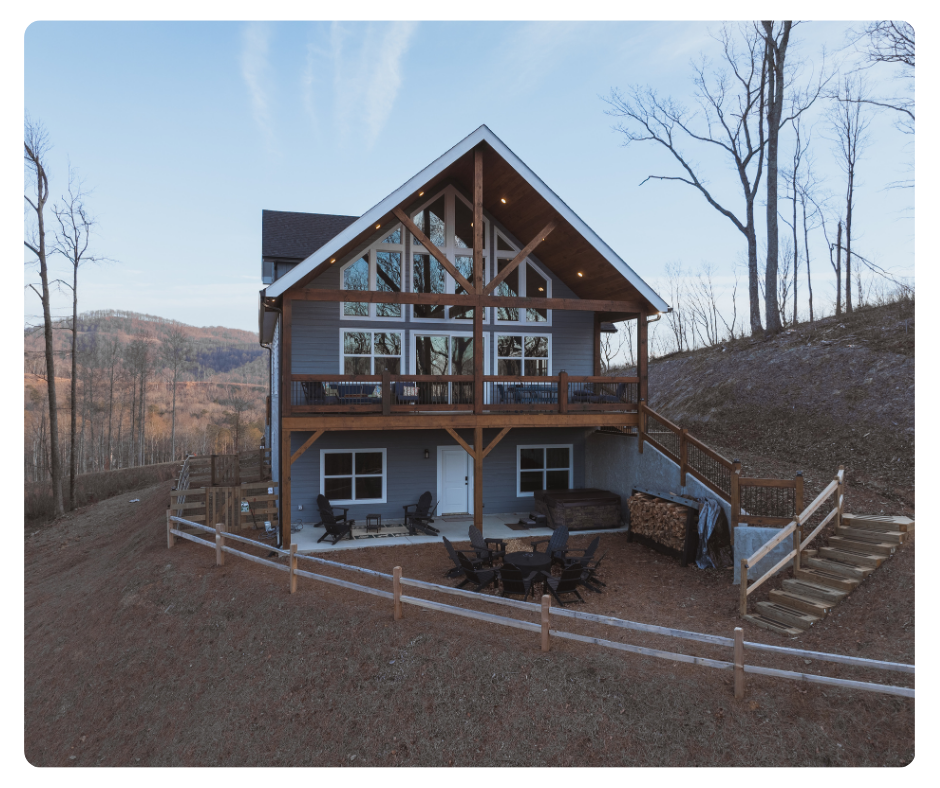 Rear view of a large mountain vacation home perched on a hill, featuring a grand timber-framed gable roof, a wall of windows, an expansive upper deck, and a lower patio with a fire pit and hot tub. Reynard Custom Homes