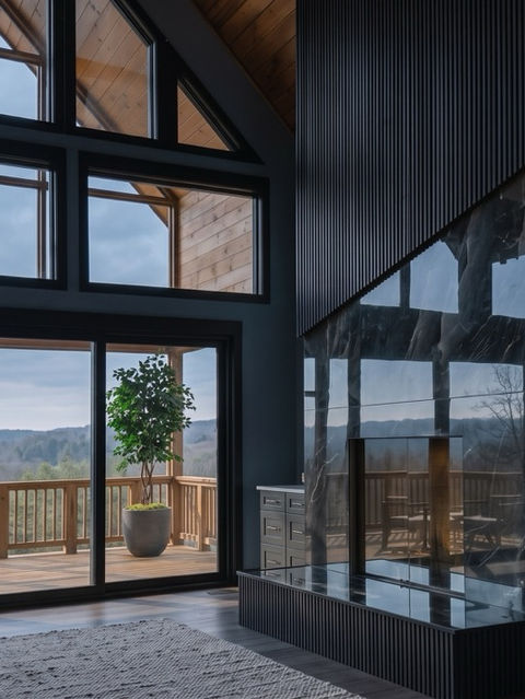 High-ceiling modern living room featuring a floor-to-ceiling black marble fireplace, dark slatted accent wall, built-in cabinets, and large glass windows overlooking a mountain view deck. Reynard Architectural Designs in Talking Rock, GA