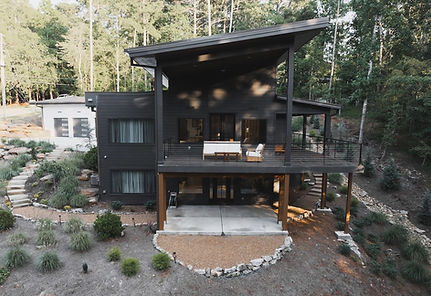 Rear elevation drone view of a mountain modern custom home in Canton, GA, showcasing a multi-level wood deck with black cable railings, a lower walk-out patio, and dramatic sloped rooflines by Reynard Architectural Designs