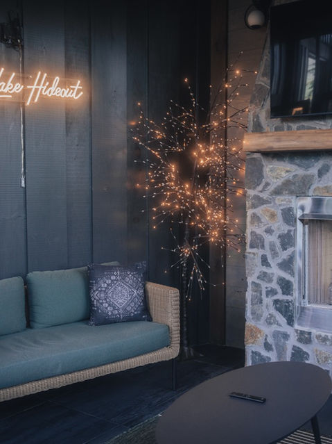 A covered outdoor patio featuring a modern sofa, a lighted decorative tree, a neon sign, and a custom stone fireplace with a fire burning inside. Architecture by James Knight Photography by Kalyn Kight. Reynard Architectural Designs