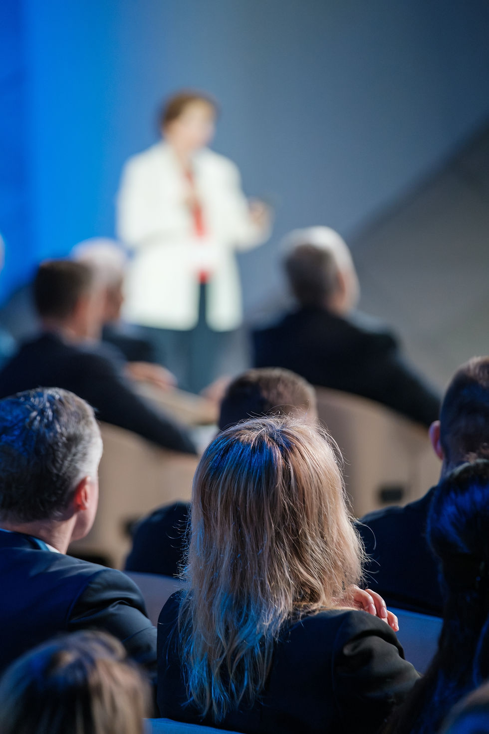 A focused image showing the back of attendees listening to a businesswoman speaking at a c