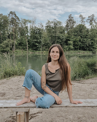 Junge Frau mit langen braunen Haaren sitzt barfuss und entspannt auf einer Holzbank am Seeufer, umgeben von Wald und Natur.
