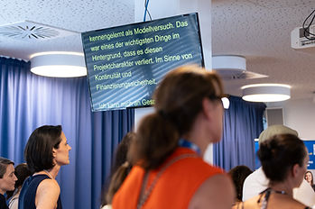 Personen im Publikum richten ihren Blick auf den Vortrag, ein Monitor an einer Säule zeigt den Text der Schriftdolmetschung.