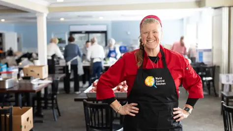 Jeni Wheeler, a member of World Central Kitchen, smiling and wearing a red chef jacket and a black apron with the World Central Kitchen logo, standing in a busy kitchen with other volunteers in the background.