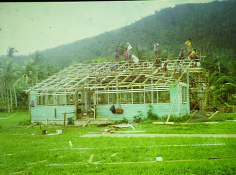 A wooden classroom building in Samoa is being renovated by students of the Don Bosco Technical Center, Samoa. In the background is a verdant green mountain.