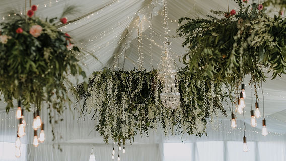 Chandelier-style hanging greenery arrangements hanging from a white wedding tent, along with white string lights and an actual chandelier.
