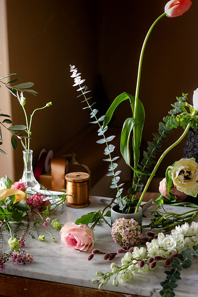 A florist work surface covered in stems of pink roses, soft pink ranunculus, white delphinium, pink wax flower, and pussy willow wish a pinkish purple tint.