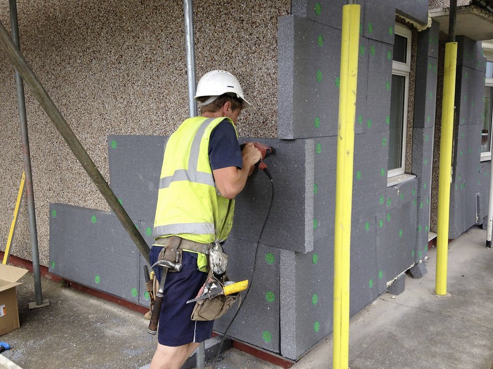 A construction worker installs gray insulation panels on a building. He wears a white helmet and yellow vest. Yellow poles are visible.