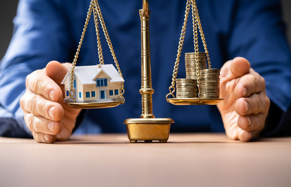 Scales balance a house model and coin stacks on a table, symbolizing financial decisions. Hands in a blue shirt adjust the scales.