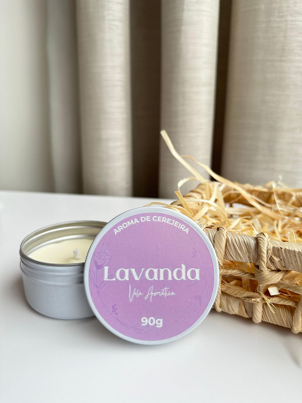 Uma vela redonda de Lavanda com 90 gramas, aberta com a tampa encostada em sua lateral próximas a uma cesta rústica com palhas dentro.