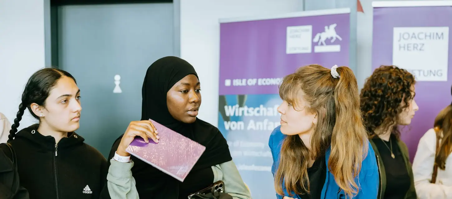 Drei junge Frauen unterhalten sich an einem Informationstisch, "Wirtschaft von A-Z" Broschüren.