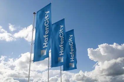 Three blue HafenCity banners waving on flagpoles against a bright blue sky with white clouds. 