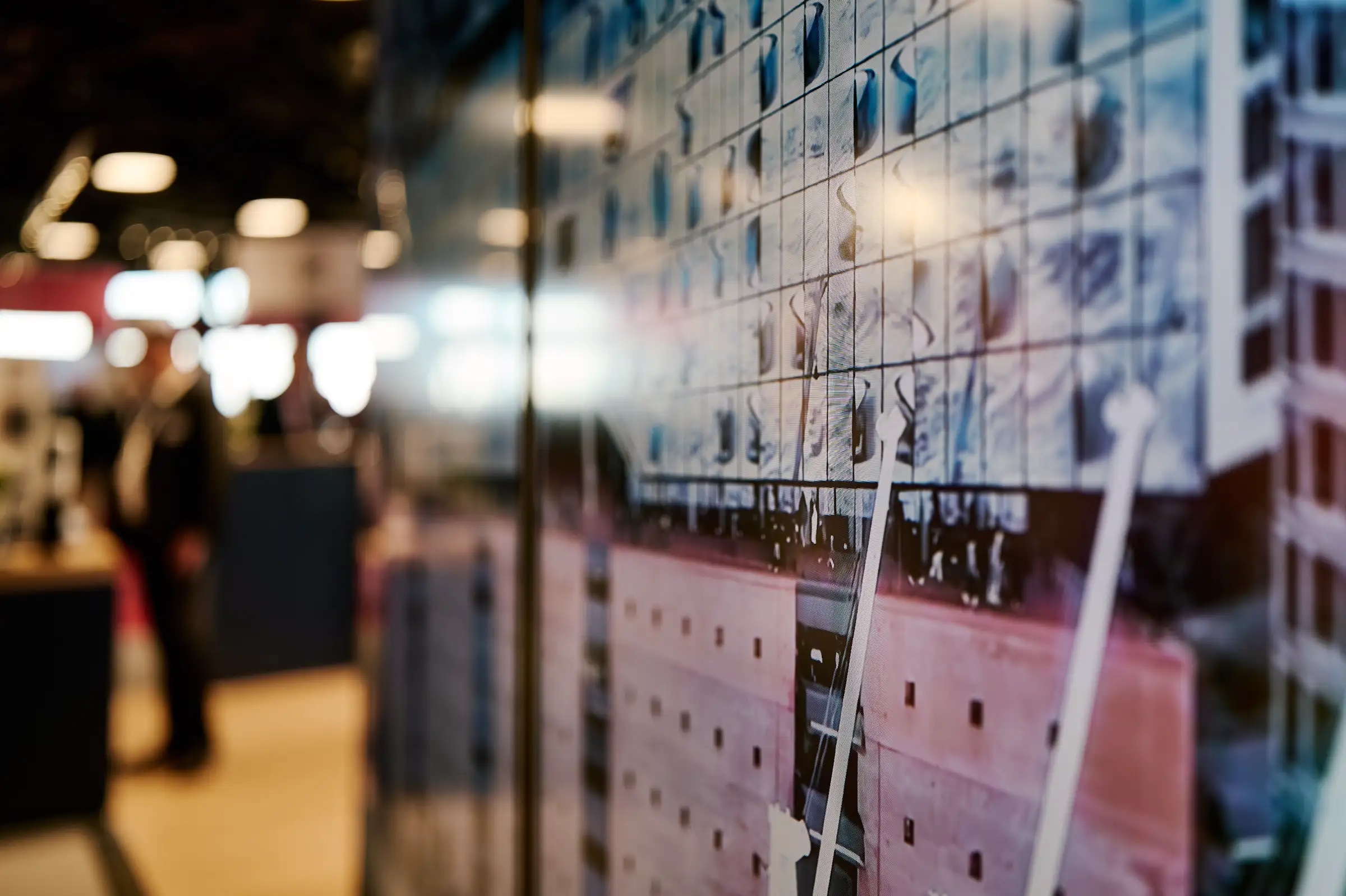 Close-up of a digital panel at a trade fair displaying architectural facade patterns, with a shallow depth of field showing a blurred event hall and visitors in the background.