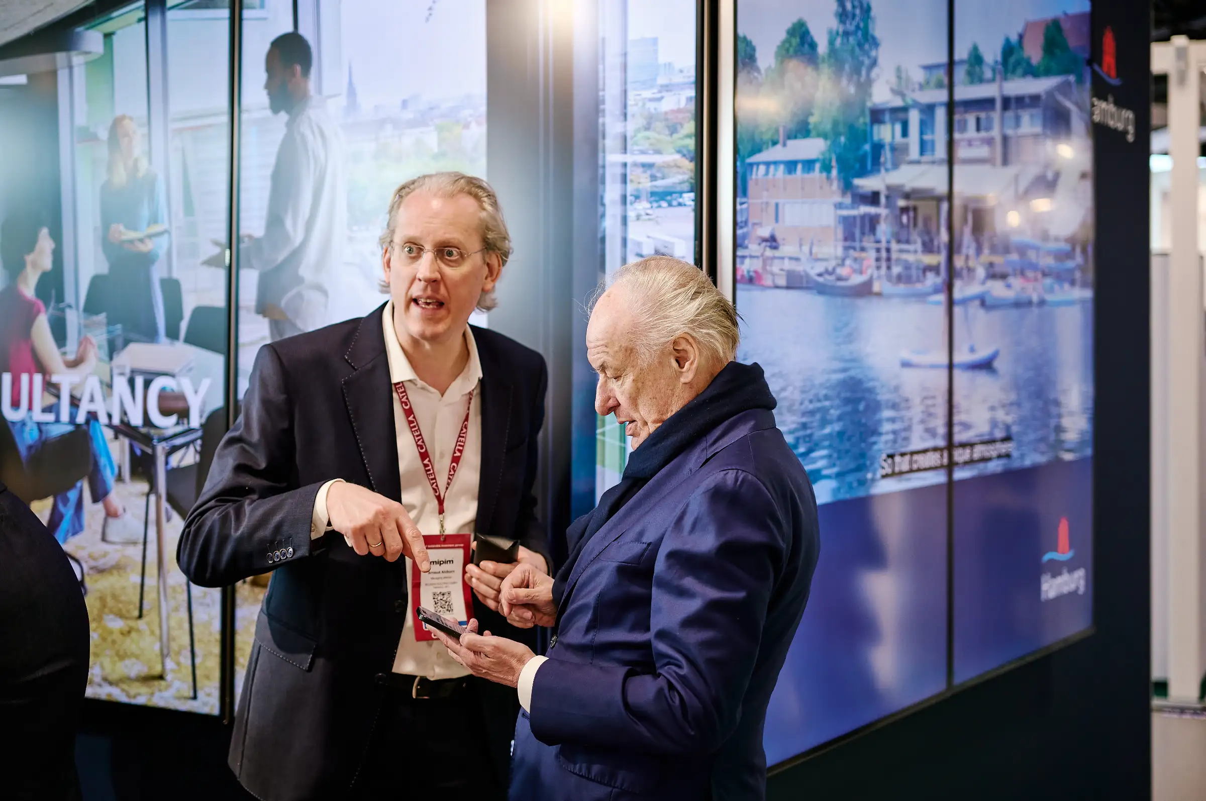 Two professionals at a trade fair discuss content on a smartphone in front of large digital screens displaying Hamburg's urban development and consultancy projects.
