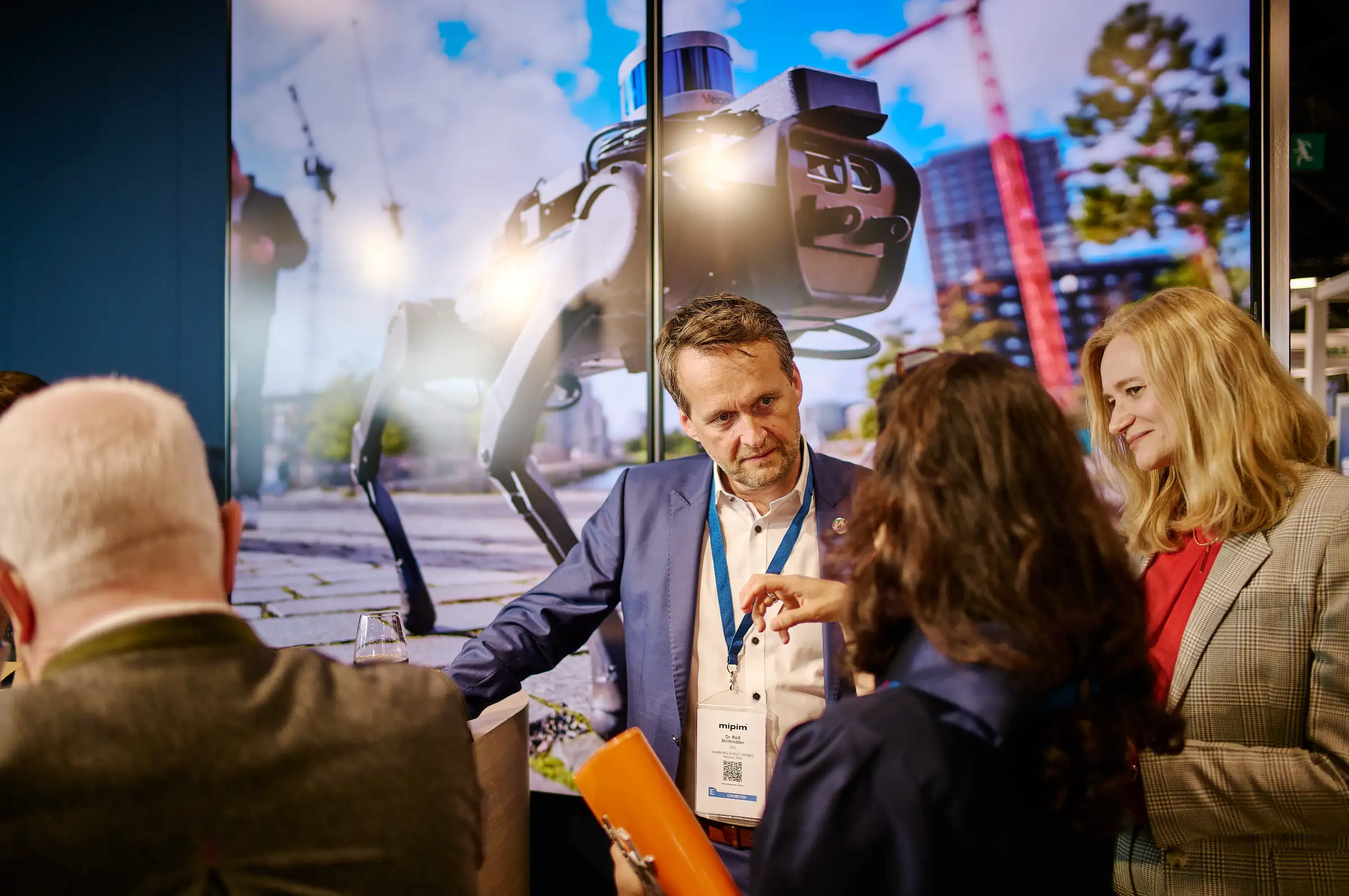 Dr. Rolf Strittmatter networking at the Hamburg stand during MIPIM 2024, in front of a large digital screen displaying a robotic dog in an urban environment.