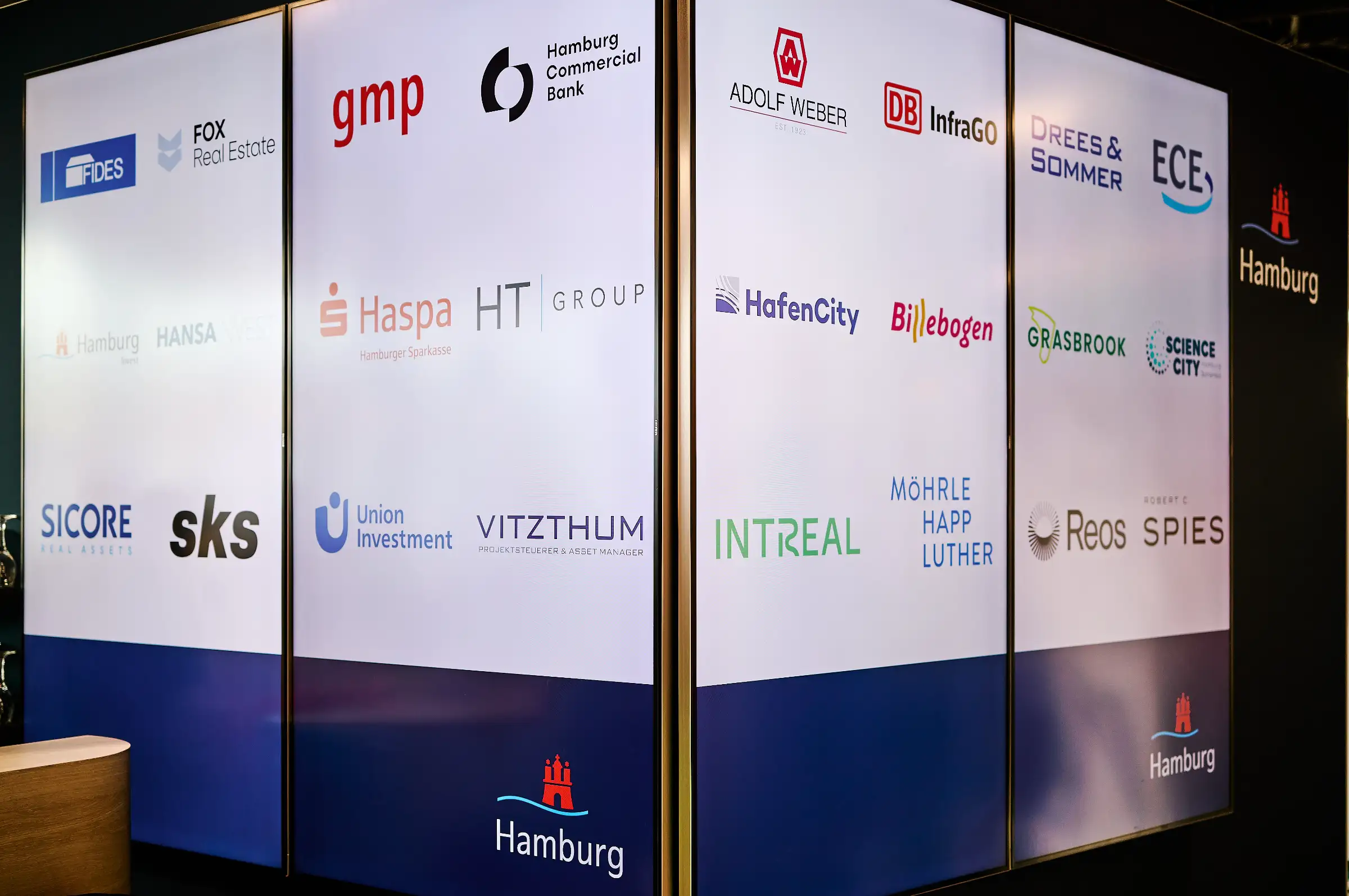 A display wall at a trade fair featuring a grid of partner logos like Haspa and HafenCity, all presented under the official Hamburg city branding on bright light panels.