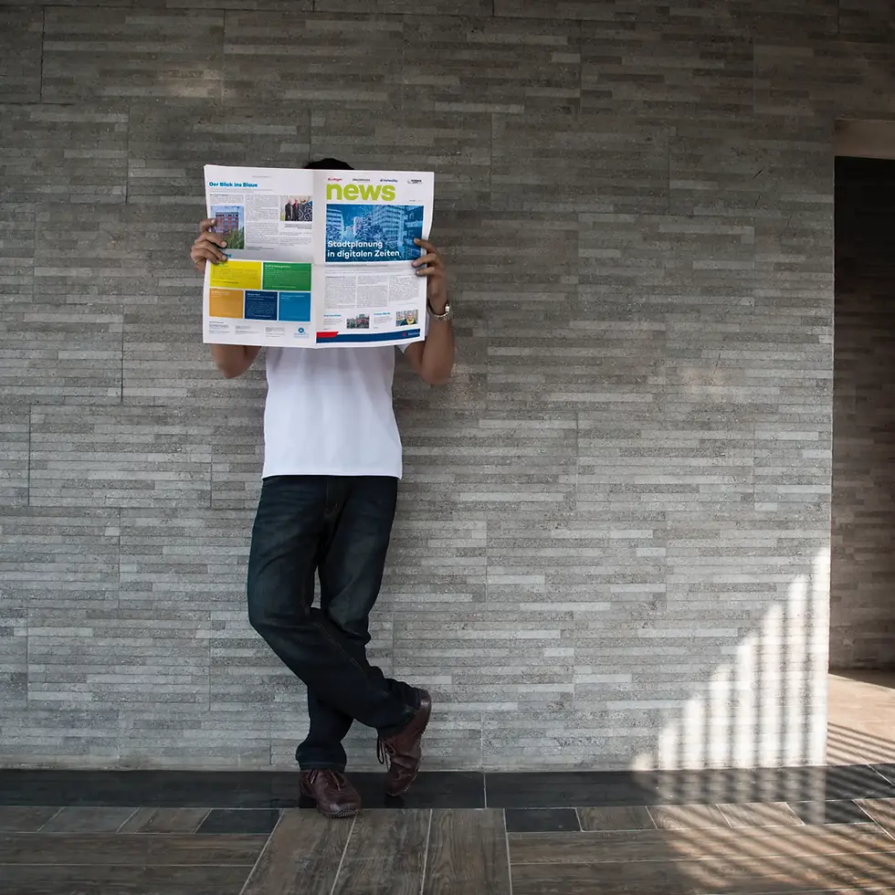Man in white shirt and jeans reads a German news magazine, leaning against a gray tiled wall with sunlit floor stripes.