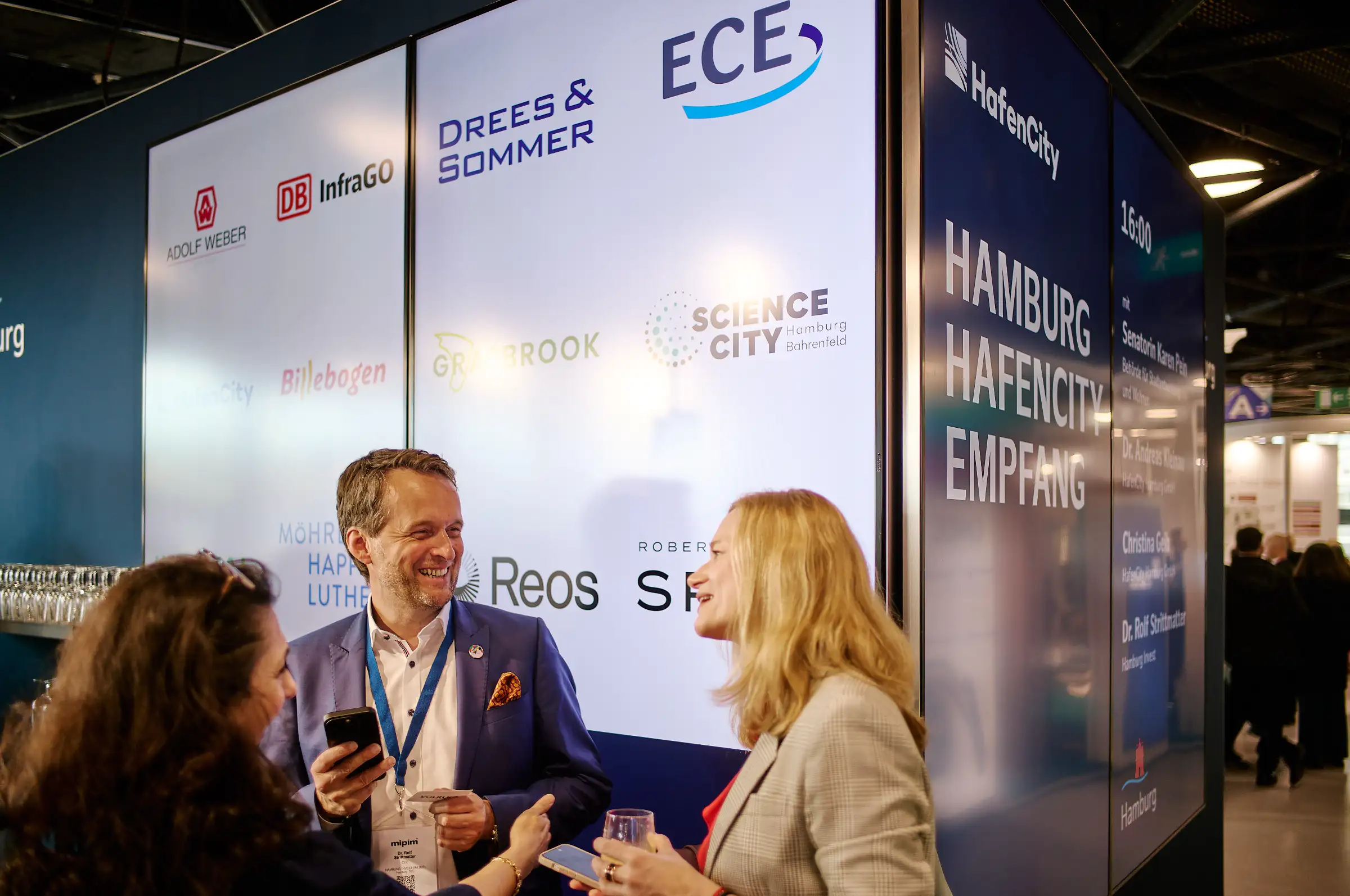 Dr. Andreas Kleinau networking with guests at the Hamburg stand during MIPIM 2024, in front of a digital display featuring partner logos and urban development projects.