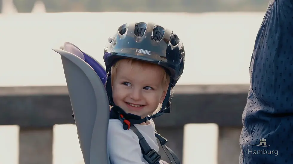 A smiling toddler with a dark bicycle helmet sitting in a child seat in front of a Hamburg waterfront.