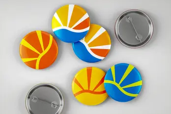 Various colorful pin-back buttons featuring the „elbsommer“ sun logo scattered on a gray surface.