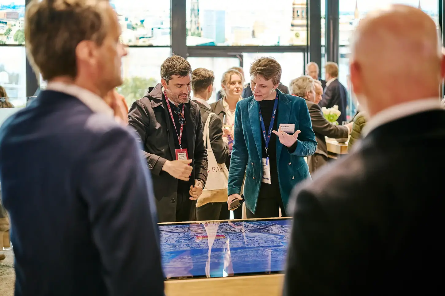 Professionals at an event interact with a large horizontal touchscreen showing a digital map of Hamburg. A man and woman look down at the display while talking.