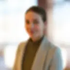 Bild von Sanja Tordini.Professionelles Porträt einer lächelnden jungen Frau mit braunem Haar, die einen grauen Blazer über einem grünen Rollkragenpullover trägt. Vor unscharfem Hintergrund.