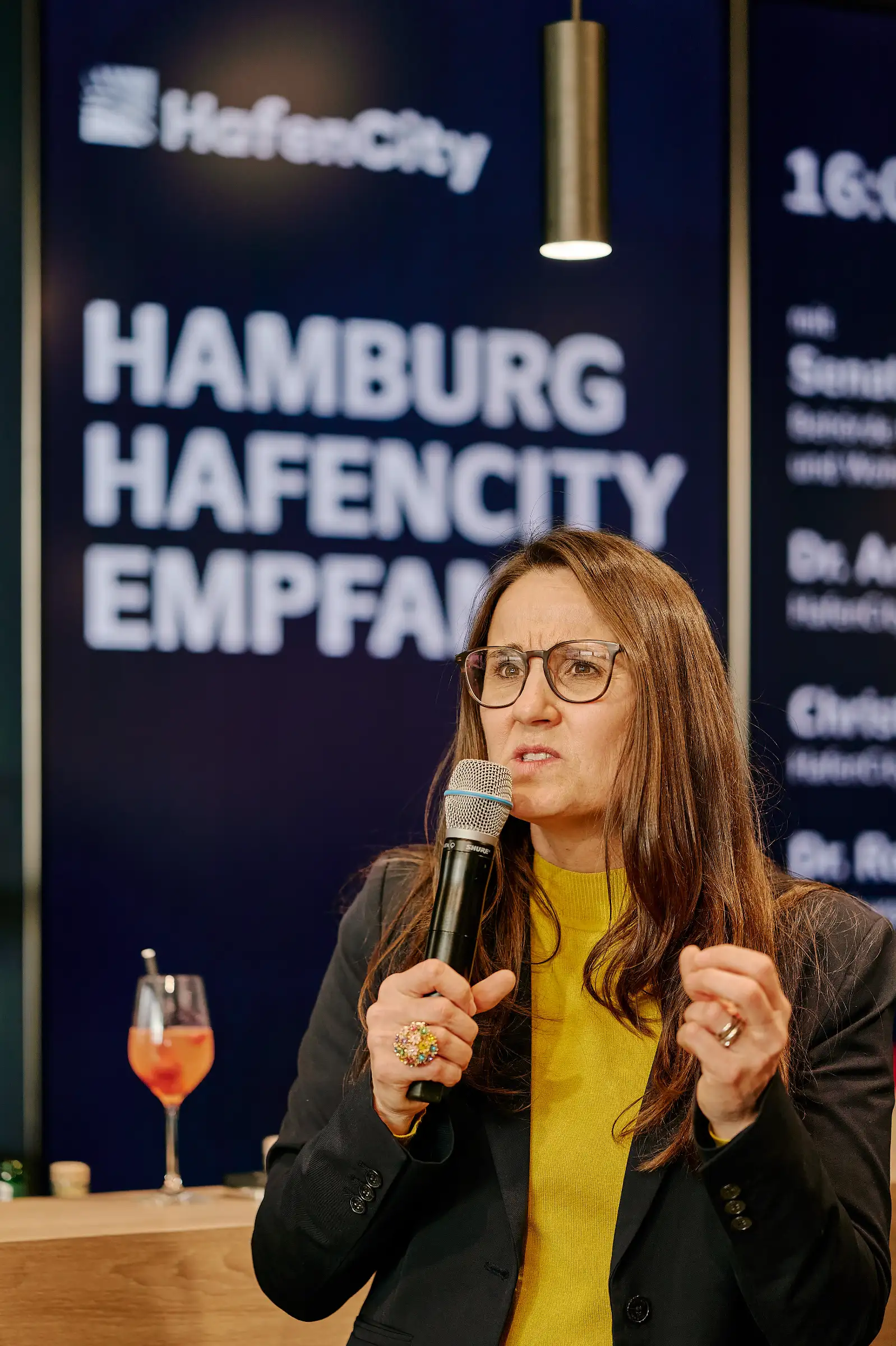 Hamburg Senator Karen Pein speaks into a microphone during the "Hamburg HafenCity Empfang" at the MIPIM trade fair, with official event signage in the background.