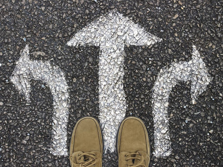 A person's shoes are seen on pavement with three arrows (left, right, and straight), indicating a choice in direction.