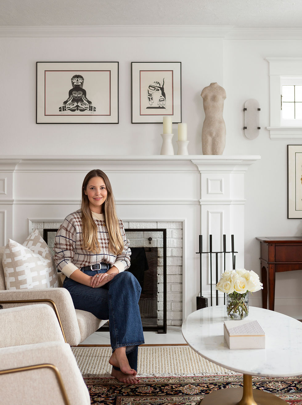 A female designer sits barefoot on a beige chair in a white living room. Art and sculptures adorn the mantel. White roses on a marble table. Cozy setting.