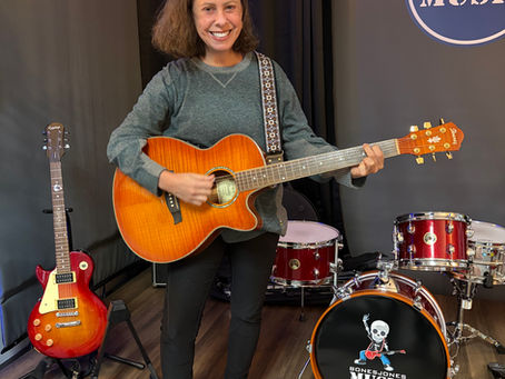 Featured student Allison Berg Karasik wears a grey shirt and black pants, smiling at the camera while standing on the BJM stage and holding an acoustic guitar