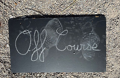 White chalk for David Deighton Dialogue Recordings art project saying Off Course in cursive on small Blackboard with footprints