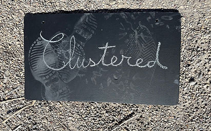 White chalk for David Deighton Dialogue Recordings art project saying Clustered in cursive on small Blackboard with footprints