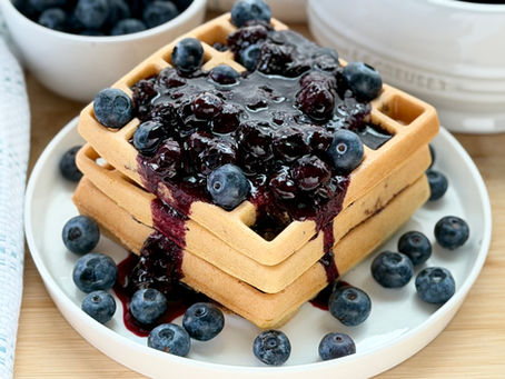 A stack of golden-brown gluten free blueberry waffles on a white plate being drizzled with thick blueberry compote and topped with fresh blueberries.