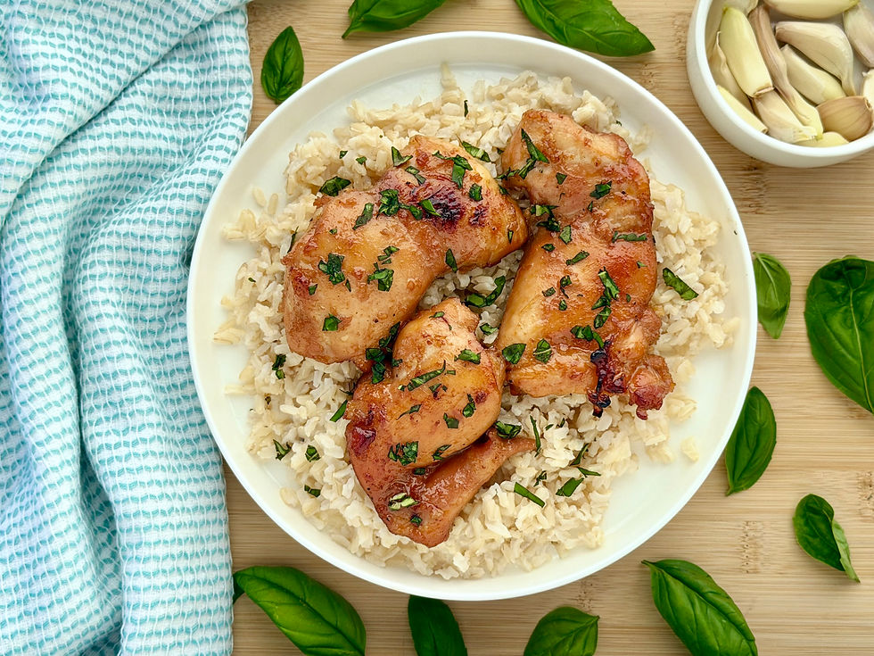 Plate of Honey Garlic Chicken Thighs over Coconut Brown Rice, topped with herbs, on a wooden board with basil leaves, garlic, and a blue checkered cloth