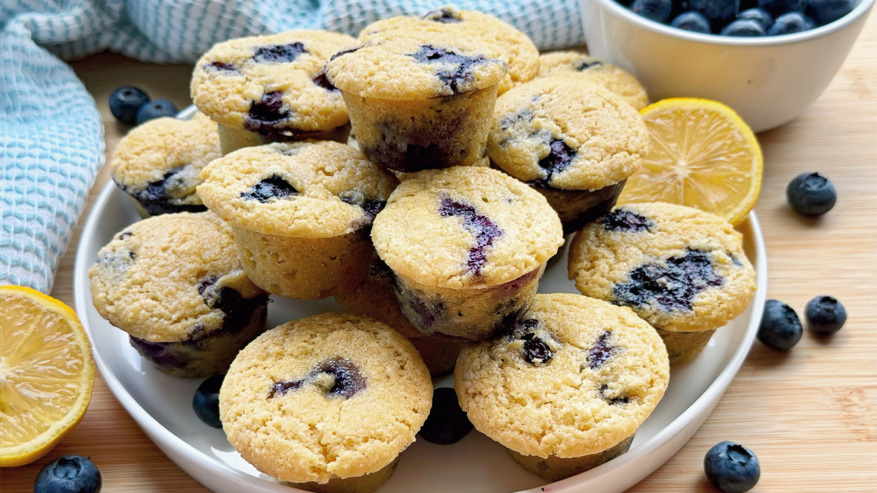 Stack of mini lemon blueberry muffins on a white plate surrounded by fresh blueberries and lemon slices