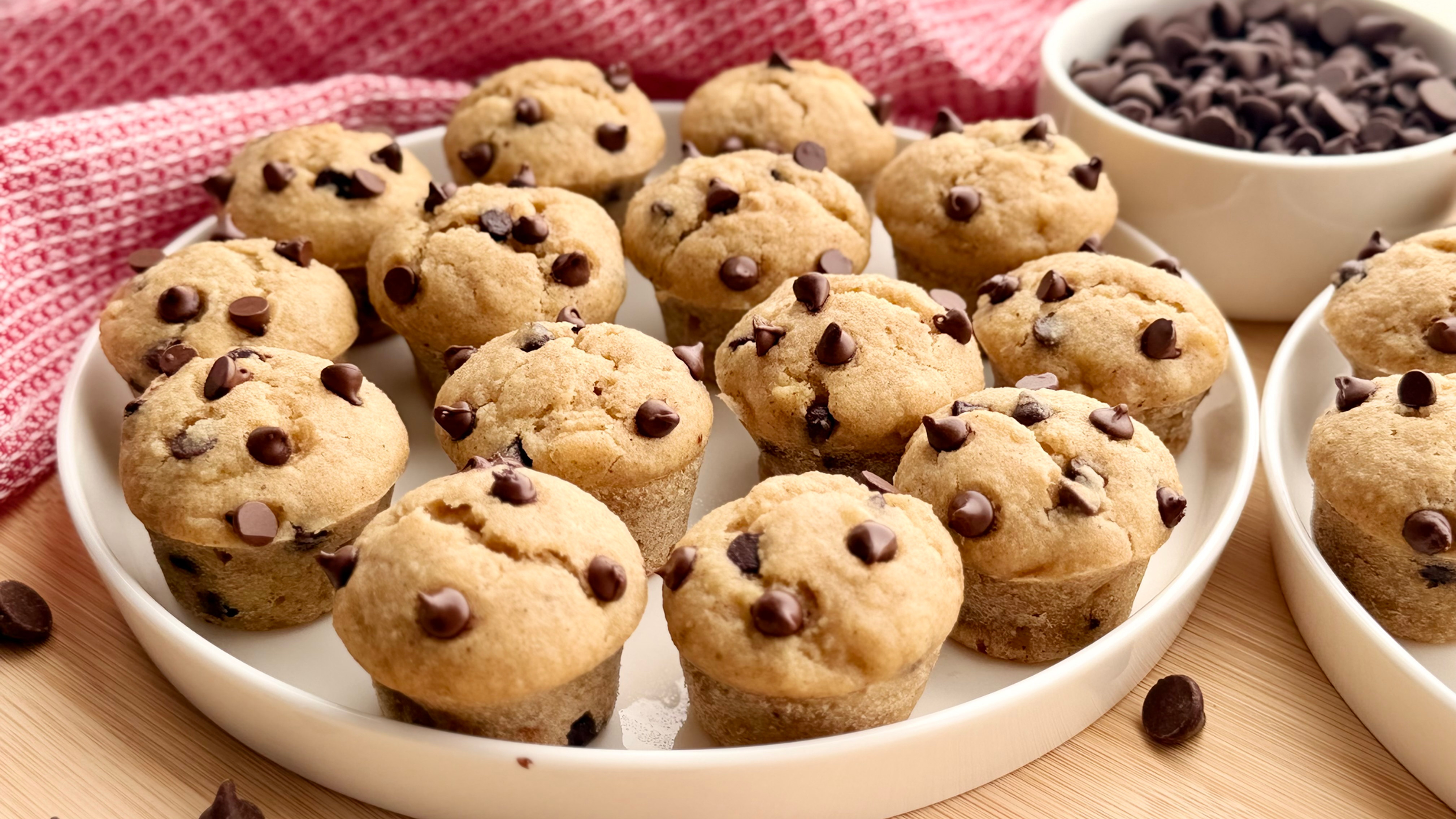 Gluten Free Banana Bread Chocolate Chip Mini Muffins on white plates, with extra chips in a bowl and scattered on a wooden table, against a red cloth.