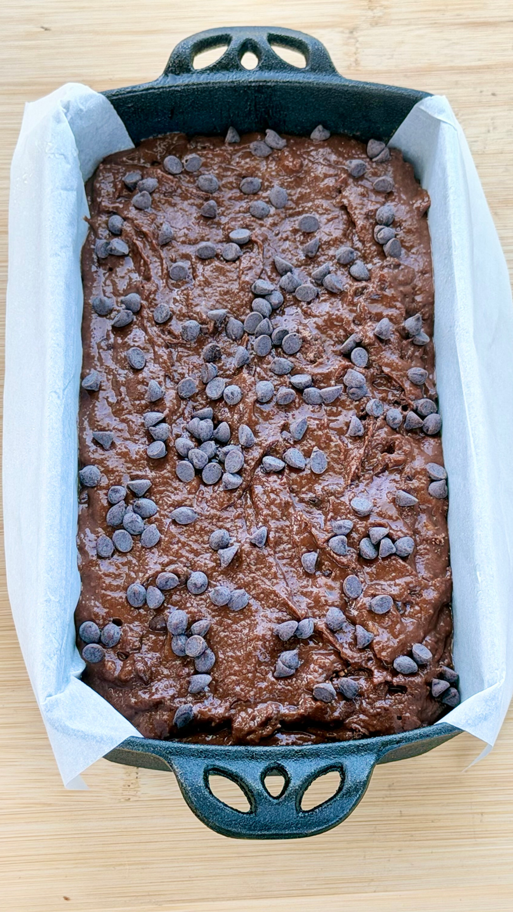 Gluten-Free Double Chocolate Banana Bread batter topped with chocolate chips in a parchment-lined loaf pan on a wooden cutting board.