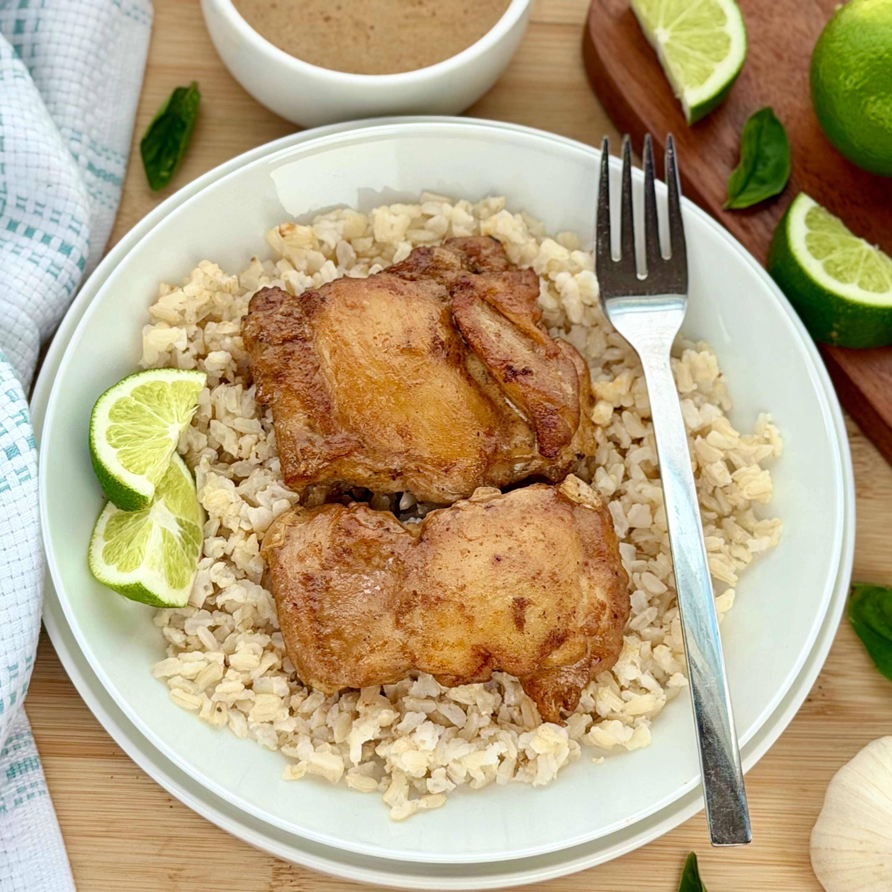 Overhead view of a serving of coconut lime chicken thighs over rice in a white bowl with a fork, lime wedges, fresh basil, and a small bowl of sauce on the side.